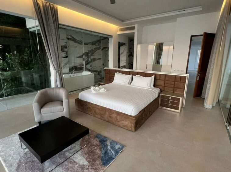 Luxurious hotel bedroom with king bed, tufted headboard, mirrored dresser, and glass-enclosed bathroom.