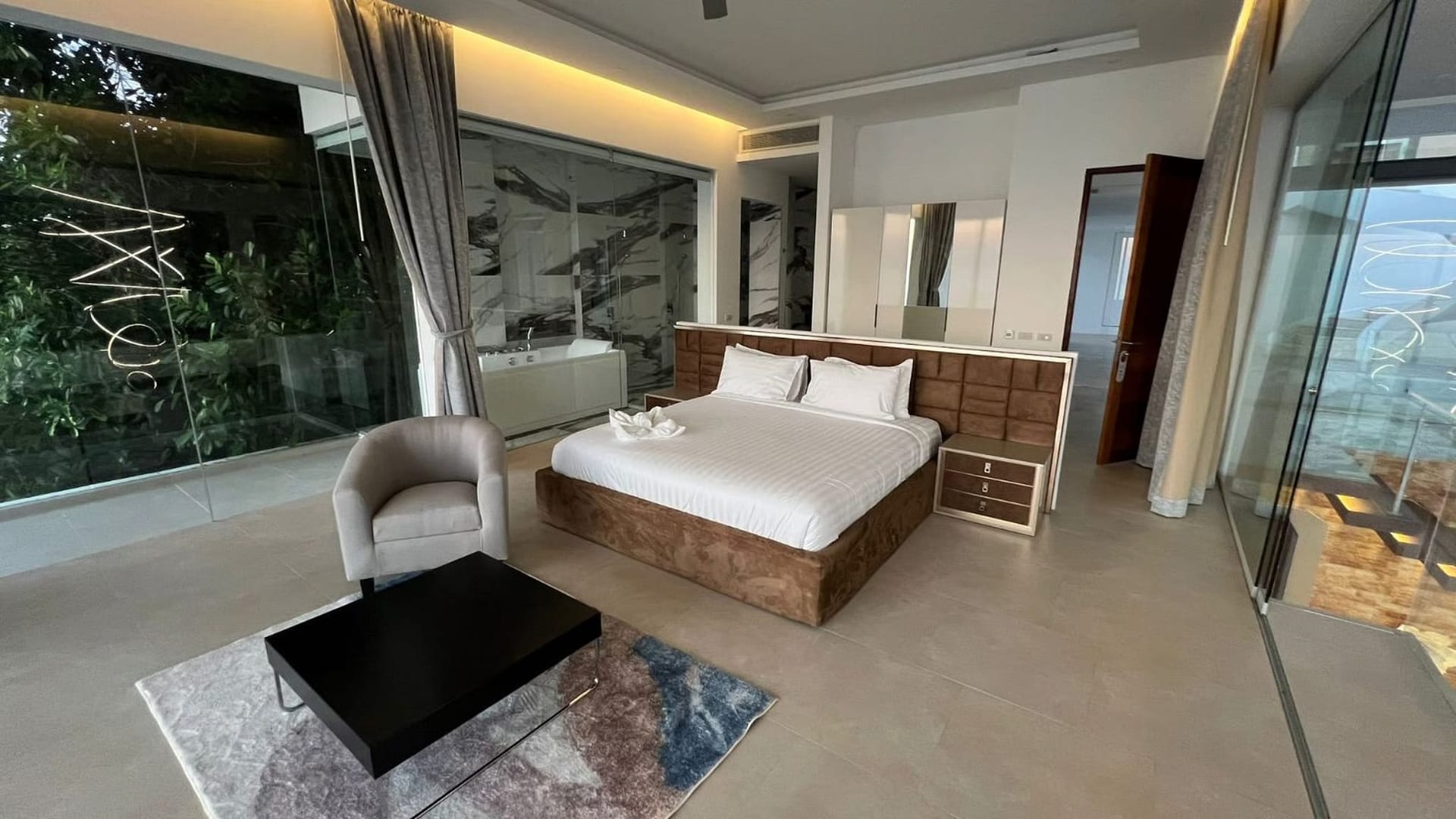 Luxurious hotel bedroom with king bed, tufted headboard, mirrored dresser, and glass-enclosed bathroom.