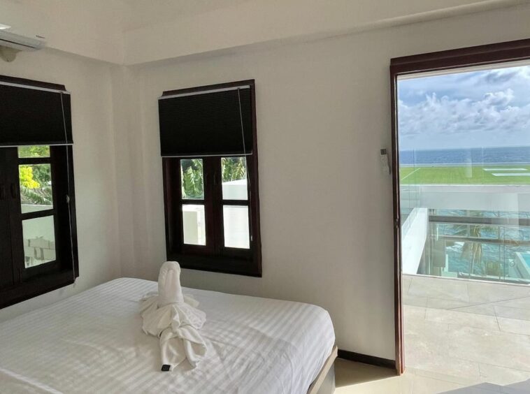 Bright hotel room with white bedding, two dark-framed windows, and a balcony door opening to an ocean view outside. Beside the bed is a towel sculpture.