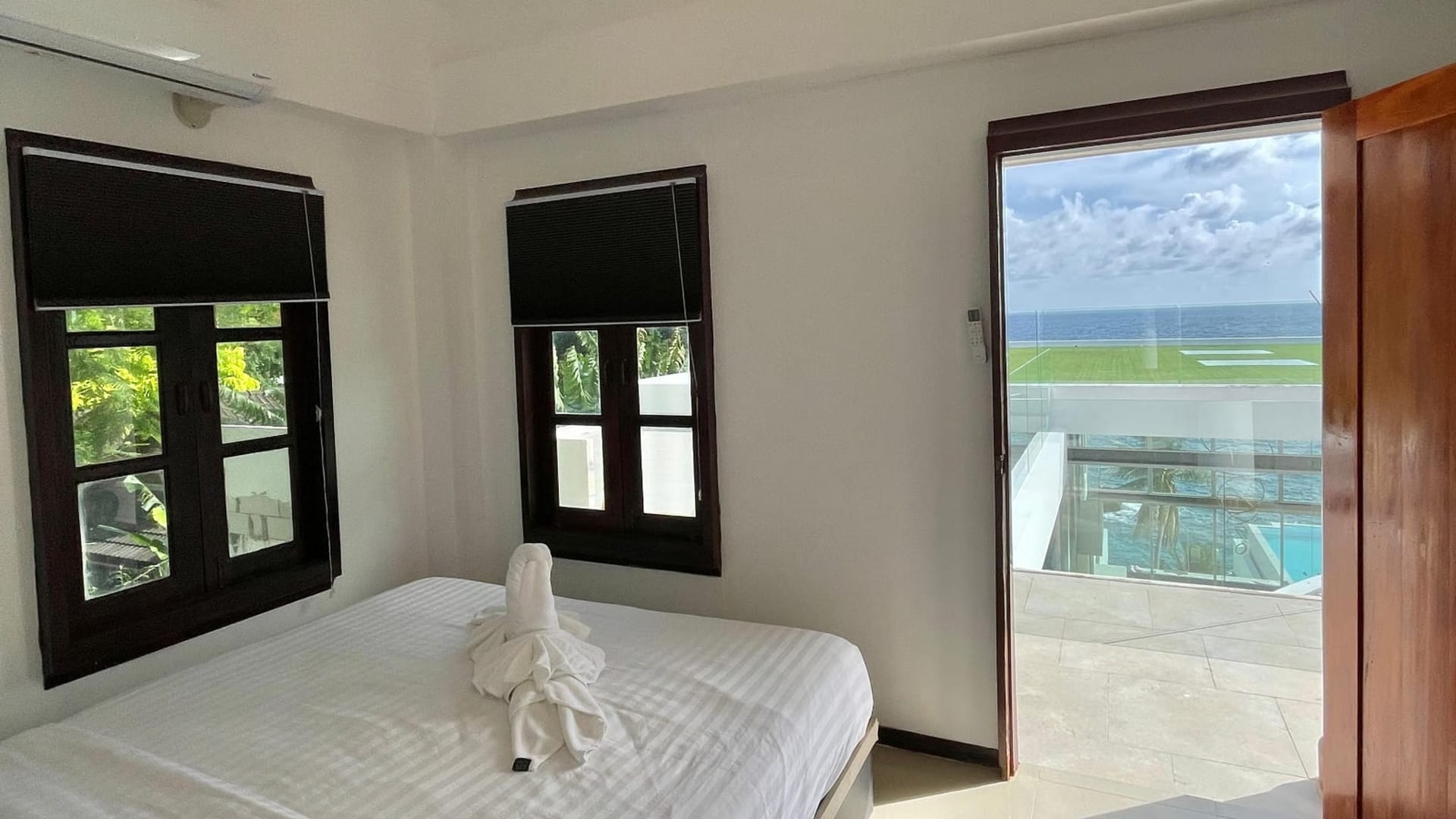 Bright hotel room with white bedding, two dark-framed windows, and a balcony door opening to an ocean view outside. Beside the bed is a towel sculpture.