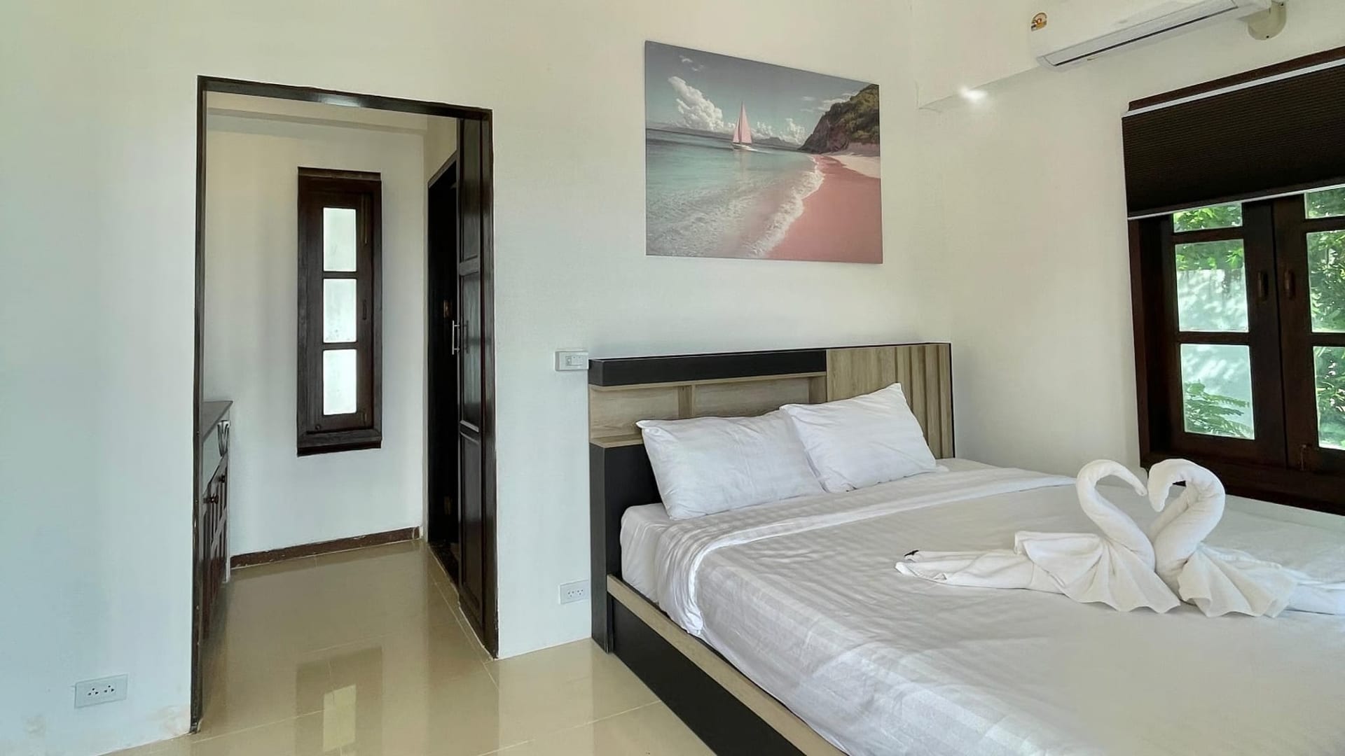 Hotel room with a large bed, white linens, and towel swans at the foot; wall art of a beach sea scene above the headboard.