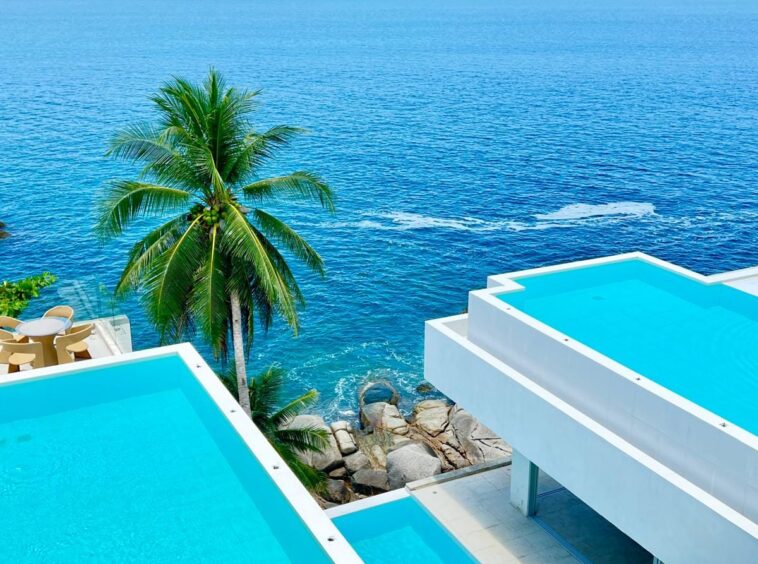 Aerial view of turquoise infinity pools at a white modern resort, with a palm tree and the blue ocean in the background.