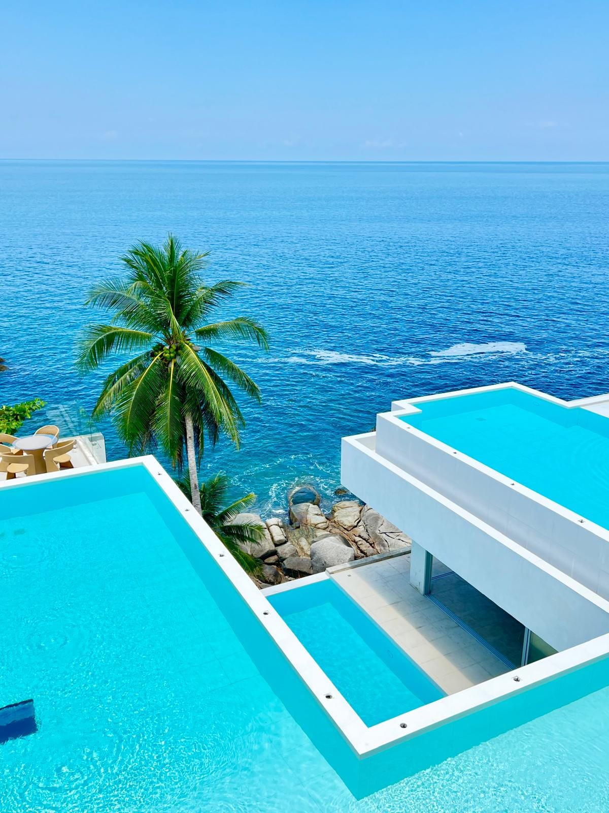 Aerial view of turquoise infinity pools at a white modern resort, with a palm tree and the blue ocean in the background.