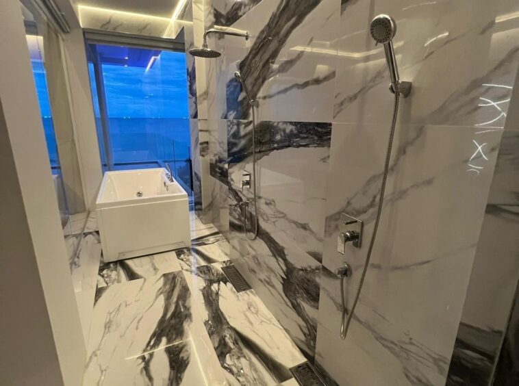 Luxurious marble bathroom with large rain shower and handheld shower fixtures, white tub near a blue-tinted window, and polished marble walls and floors.