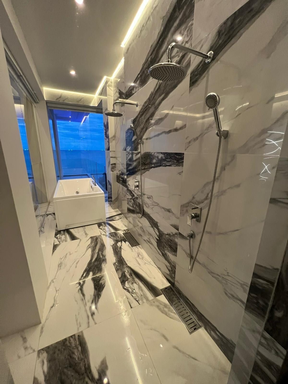 Luxurious marble bathroom with large rain shower and handheld shower fixtures, white tub near a blue-tinted window, and polished marble walls and floors.