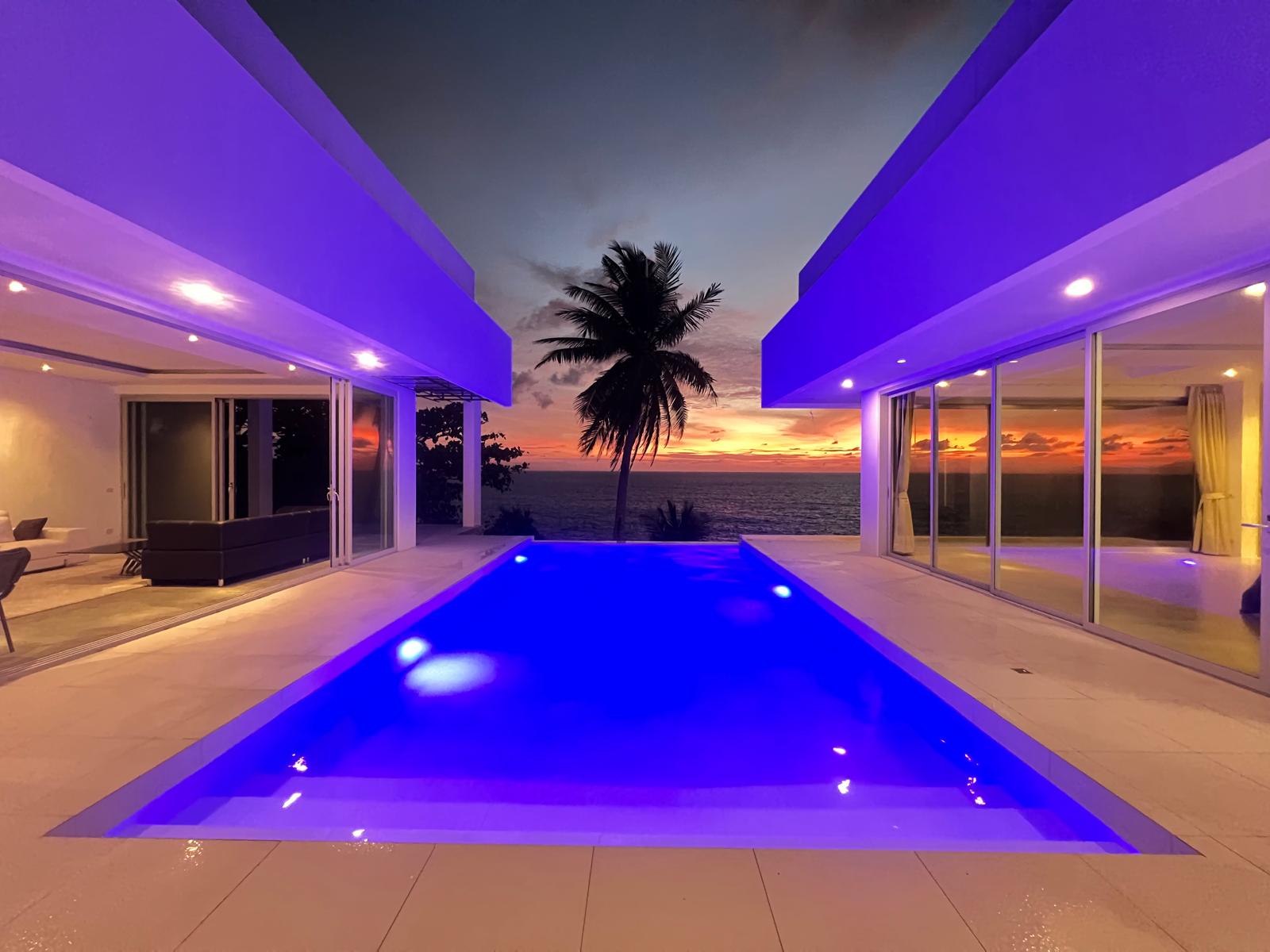 Sunset over the ocean viewed from a modern villa with a blue-lit pool between open living spaces.