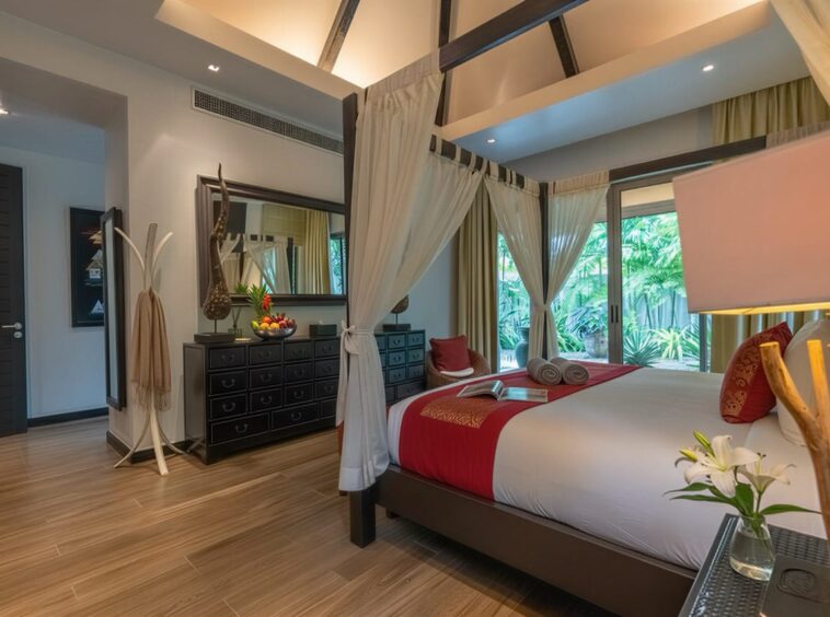 Luxurious four-poster bedroom with white drapes, red bed runner, and a view of tropical plants outside the glass doors.