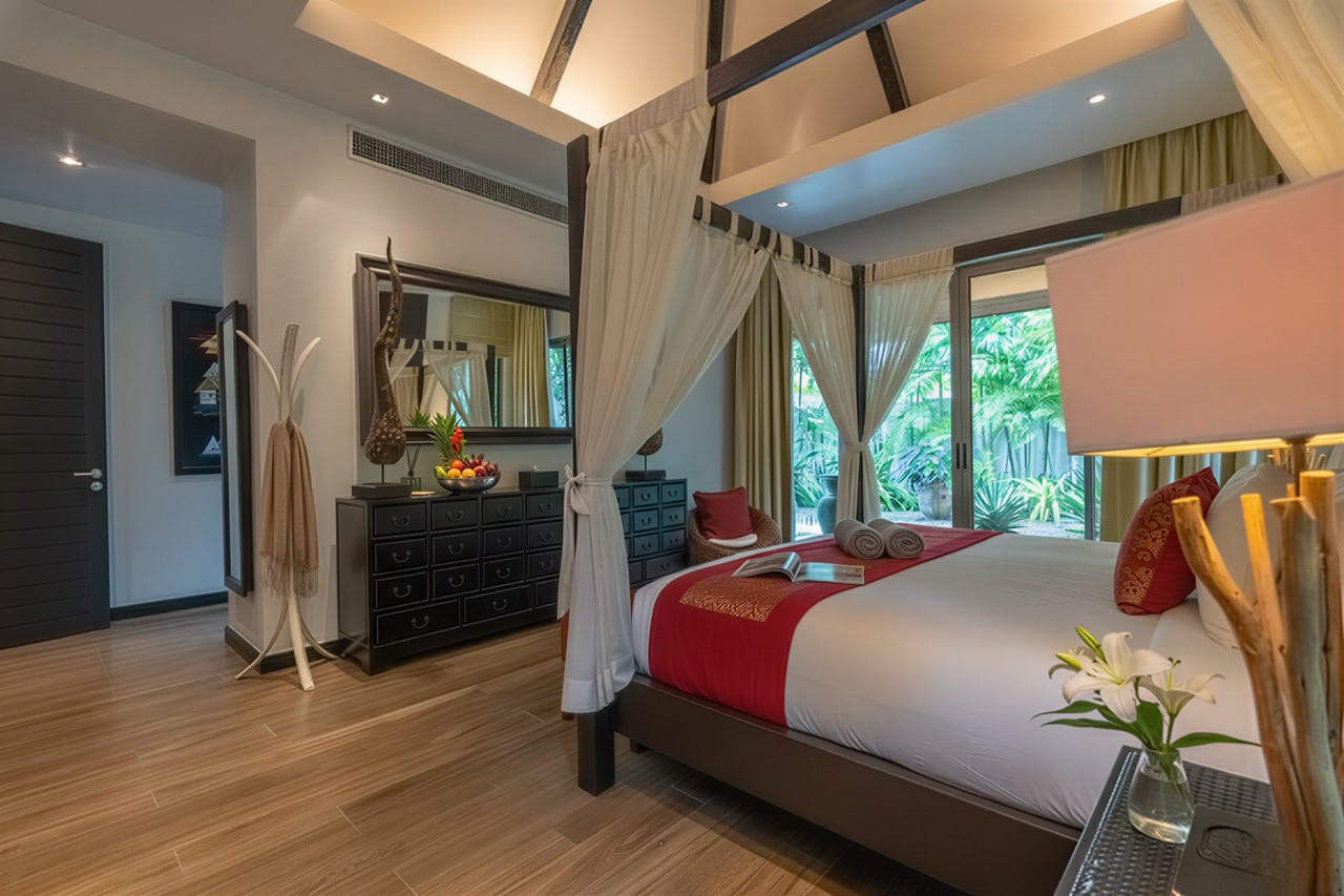 Luxurious four-poster bedroom with white drapes, red bed runner, and a view of tropical plants outside the glass doors.