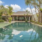 Luxurious tropical villa with a rectangular pool, palm trees, and a shaded outdoor dining area under a wooden pavilion.