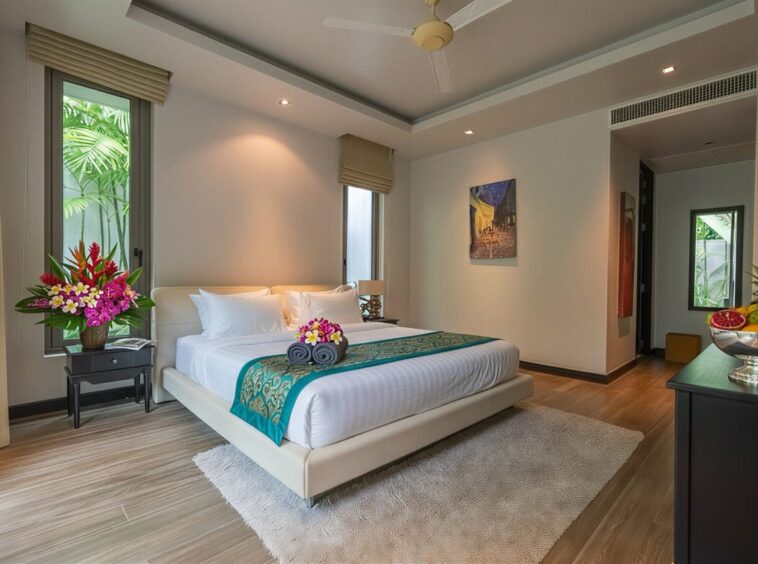 Luxurious hotel bedroom with a white platform bed, teal runner, and fresh flower arrangements on each side table; tropical plants outside the windows.