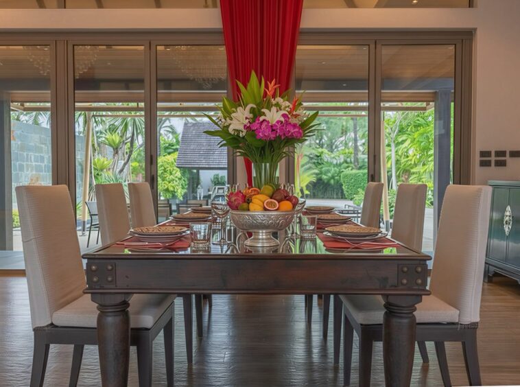 Elegant dining room with a dark wood table set for eight, white upholstered chairs, and a centerpiece of fruit and tropical flowers.