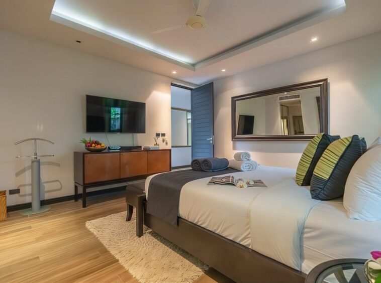 Modern hotel bedroom with a large bed, white linens, gray throw, and decorative green-and-black pillows facing a wall-mounted TV.