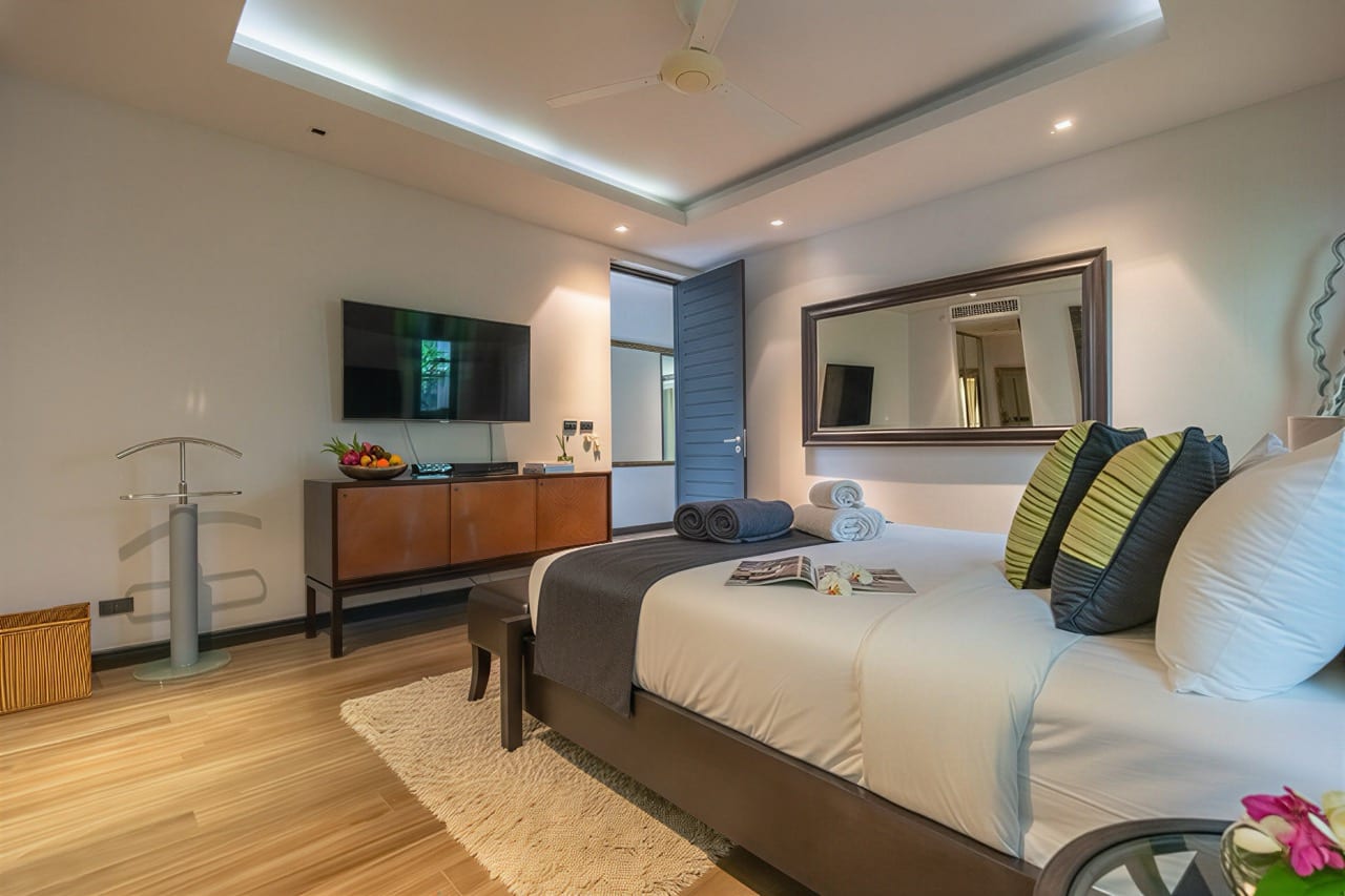 Modern hotel bedroom with a large bed, white linens, gray throw, and decorative green-and-black pillows facing a wall-mounted TV.