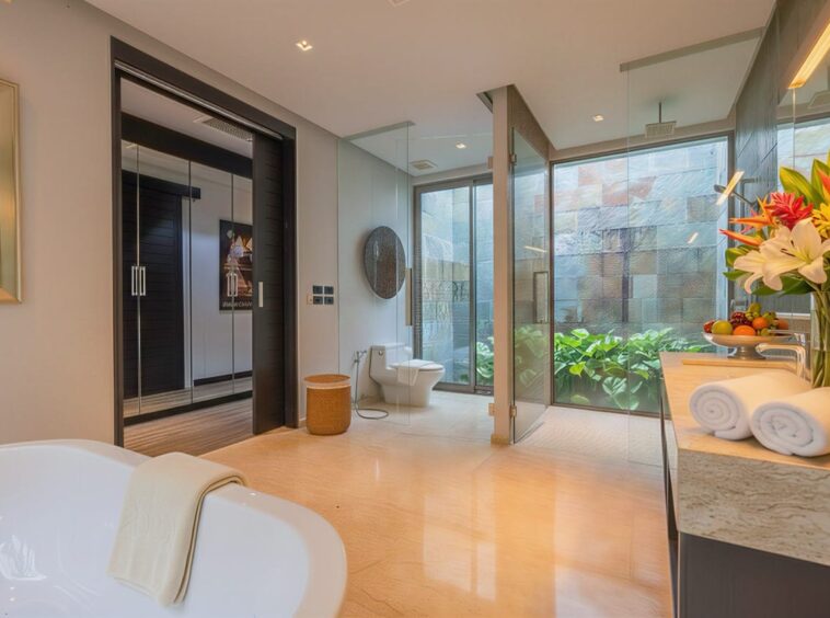 Luxurious bathroom with a freestanding tub, glass-enclosed shower, marble vanity, and a vase of lilies by a fruit centerpiece near large windows with greenery outside.