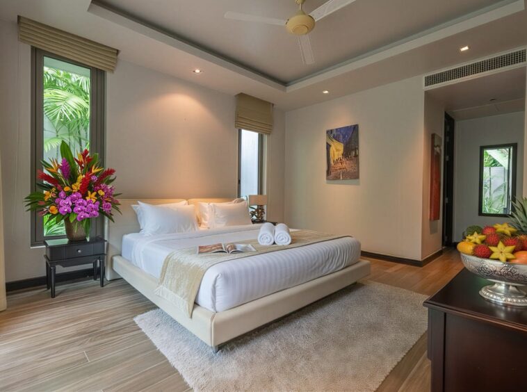 Luxurious hotel bedroom with a king bed, white linens, beige headboard, and a vase of tropical flowers on a side table beside a window. A fruit bowl sits on a dark dresser to the right. Hallway and artwork visible in the background.