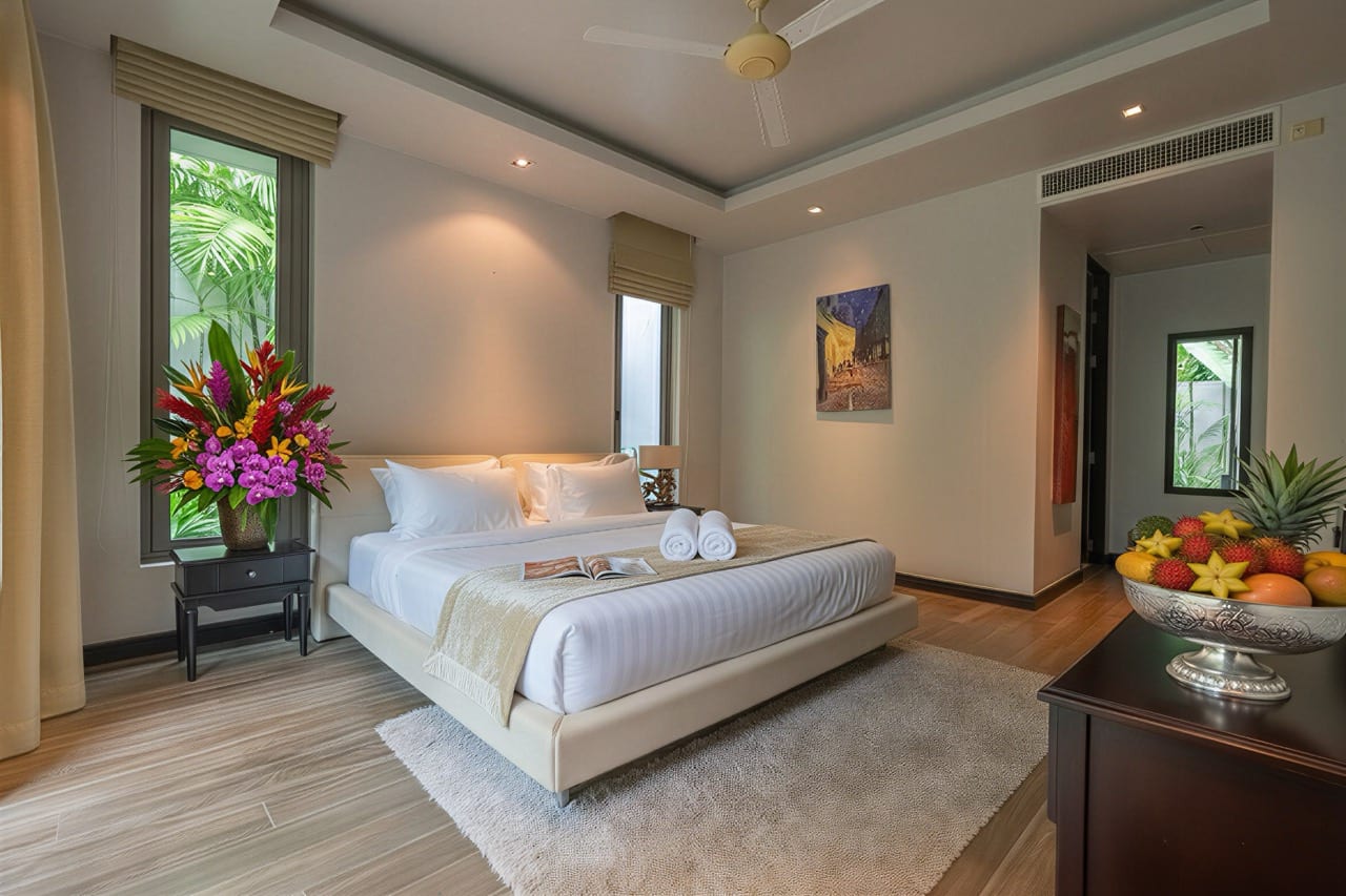 Luxurious hotel bedroom with a king bed, white linens, beige headboard, and a vase of tropical flowers on a side table beside a window. A fruit bowl sits on a dark dresser to the right. Hallway and artwork visible in the background.