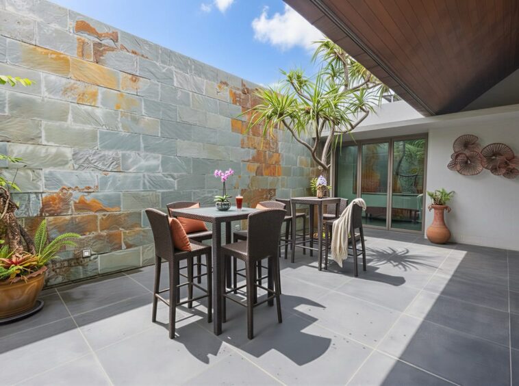Outdoor patio with two high tables and wicker chairs, set against a slate stone wall and potted greenery, near glass doors.