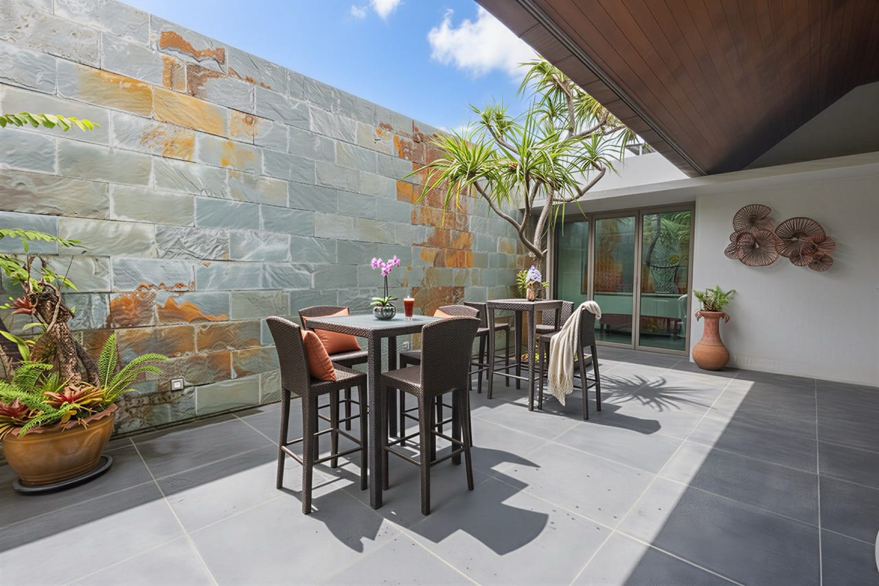Outdoor patio with two high tables and wicker chairs, set against a slate stone wall and potted greenery, near glass doors.
