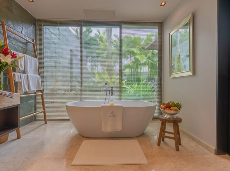 Spa-style bathroom with a freestanding tub beside large windows and lush greenery outside.