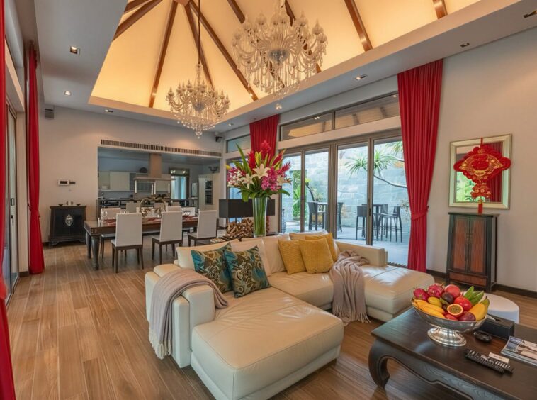 Open-concept living and dining room with a white sectional sofa, a large dining table, red curtains, and crystal chandeliers overhead, plus a fruit bowl on the coffee table near a glass door to an outdoor area.