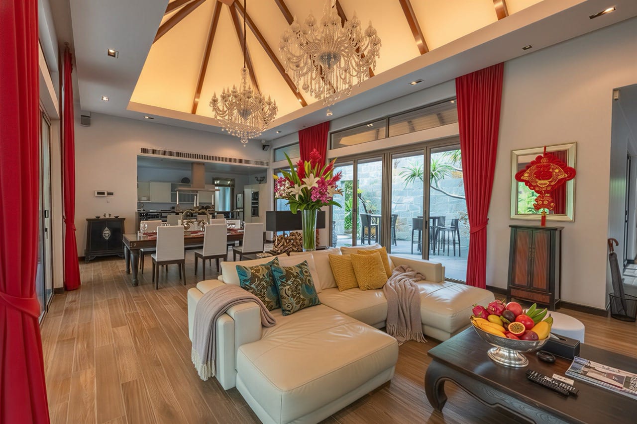 Open-concept living and dining room with a white sectional sofa, a large dining table, red curtains, and crystal chandeliers overhead, plus a fruit bowl on the coffee table near a glass door to an outdoor area.