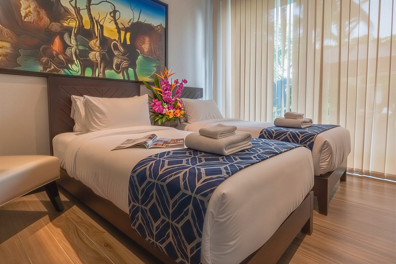 Twin hotel beds with white linens and blue geometric runners; towels, a magazine, and a floral arrangement between the headboards.