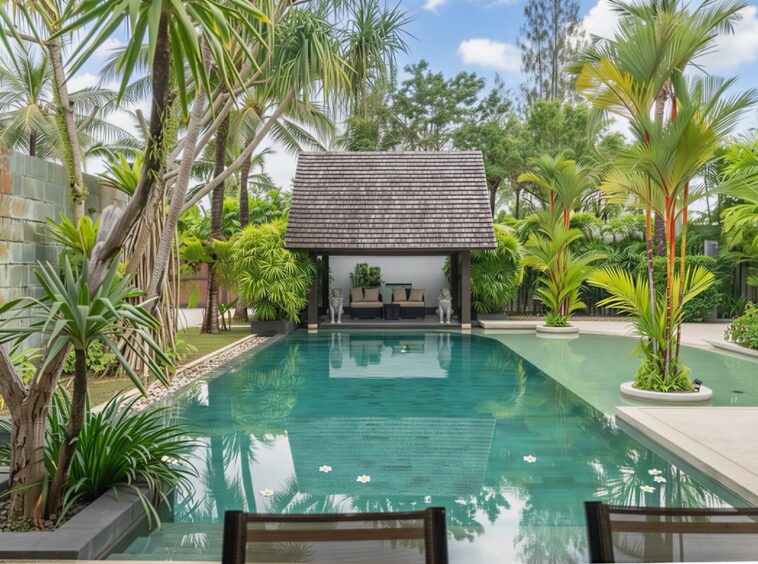 Luxurious backyard pool with palm trees, clear water, and a thatched-roof cabana with lounge seating at the far end.