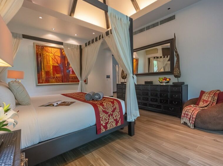 Four-poster bed with white linens and a red-gold decorative runner in a bright hotel bedroom with sheer curtains.