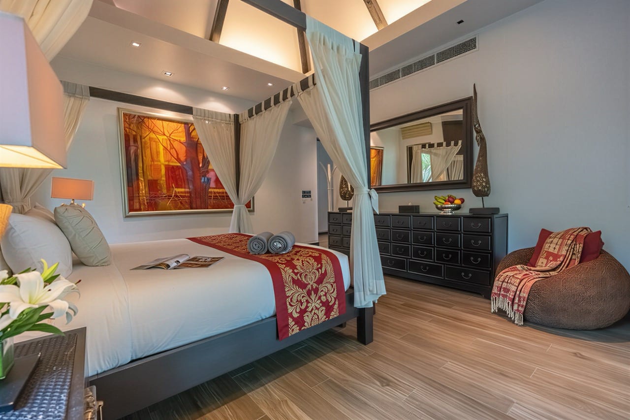 Four-poster bed with white linens and a red-gold decorative runner in a bright hotel bedroom with sheer curtains.