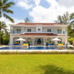 White two-story villa with red-tiled roof, pool in front, blue lounge chairs and white umbrellas set on a sunny tropical lawn.