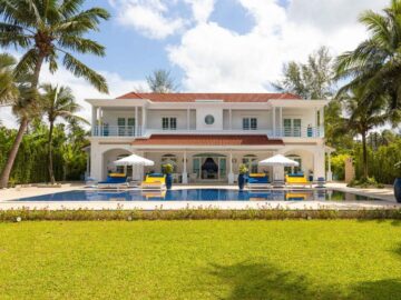 White two-story villa with red-tiled roof, pool in front, blue lounge chairs and white umbrellas set on a sunny tropical lawn.
