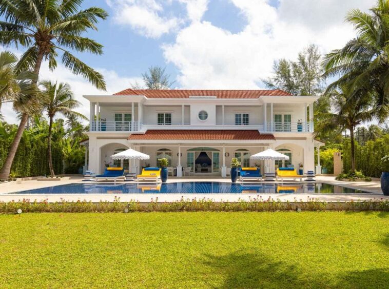 White two-story villa with red-tiled roof, pool in front, blue lounge chairs and white umbrellas set on a sunny tropical lawn.