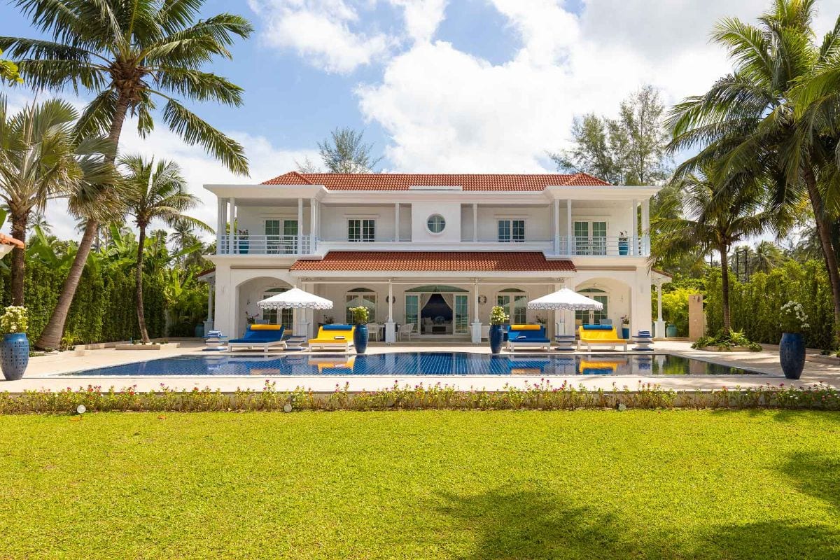 White two-story villa with red-tiled roof, pool in front, blue lounge chairs and white umbrellas set on a sunny tropical lawn.