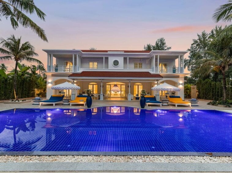 Luxurious two-story villa with a red-tiled roof and a large blue-tiled pool in the foreground at sunset.