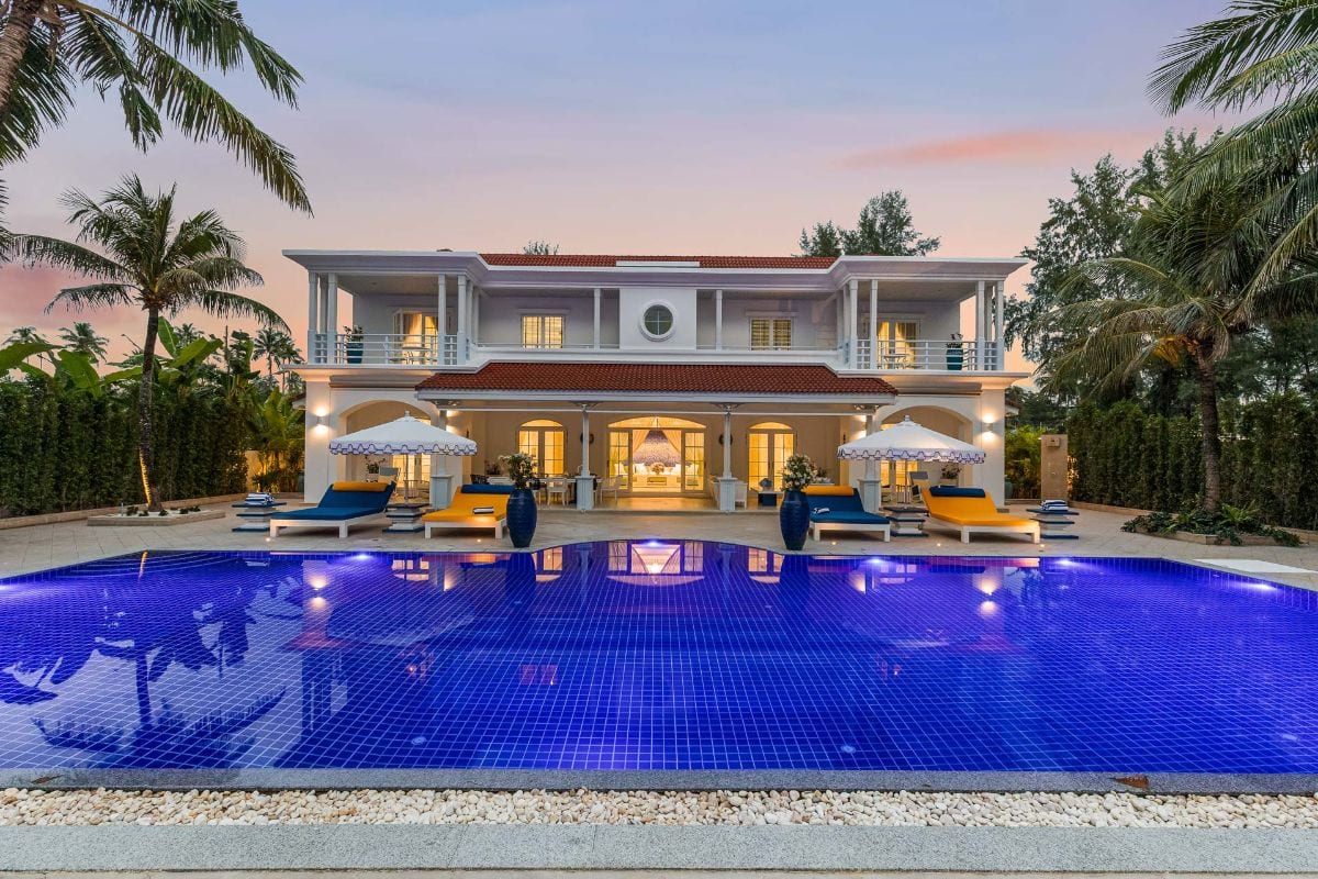 Luxurious two-story villa with a red-tiled roof and a large blue-tiled pool in the foreground at sunset.
