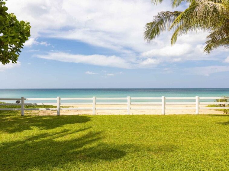A sunny beach scene behind a white fence, with green grass in the foreground and calm turquoise ocean under a blue sky with palm trees.