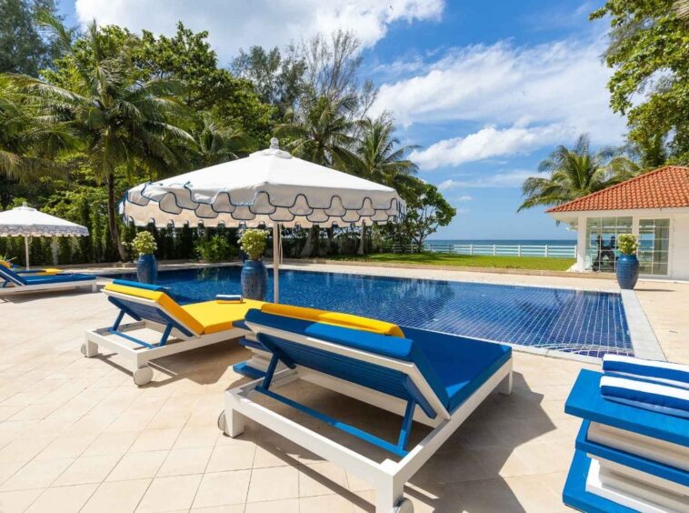 Poolside at a tropical resort: white chaise lounges with blue frames and yellow cushions under large white umbrellas, with the ocean in the distance.