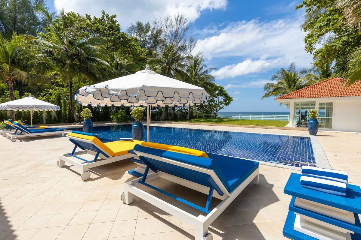 Poolside at a tropical resort: white chaise lounges with blue frames and yellow cushions under large white umbrellas, with the ocean in the distance.