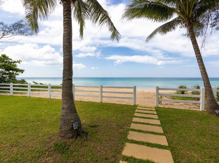 Path of stepping stones across a grassy yard toward a sandy beach and calm sea, with palm trees and a white fence on either side.