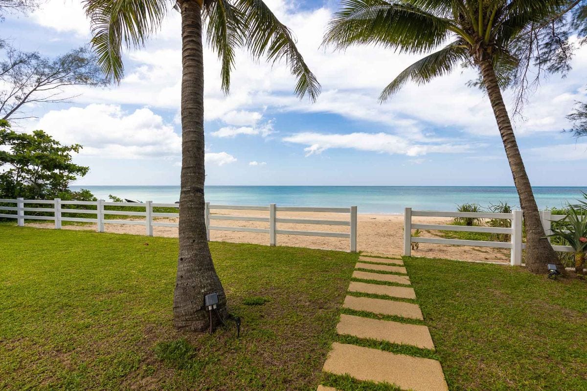 Path of stepping stones across a grassy yard toward a sandy beach and calm sea, with palm trees and a white fence on either side.