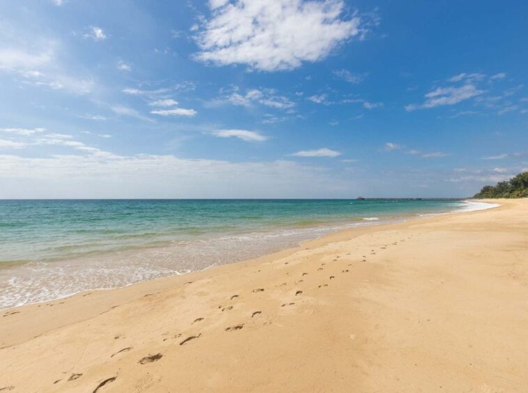 Sunny beach with turquoise water, blue sky, and footprints in the wet sand leading toward the water.