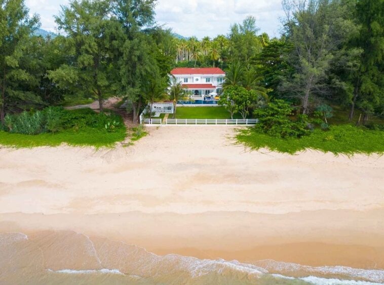 Aerial view of a white two-story house with a red roof nestled among dense green trees, a white fence in front, overlooking a wide sandy beach.