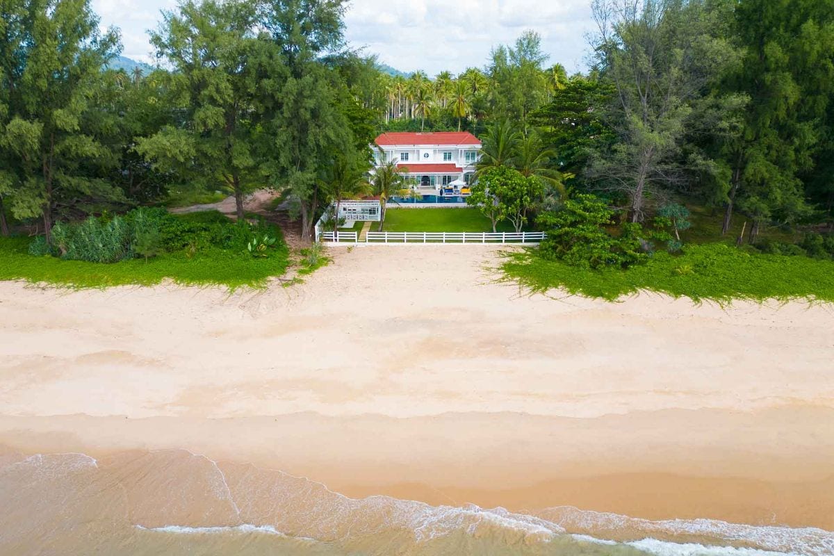 Aerial view of a white two-story house with a red roof nestled among dense green trees, a white fence in front, overlooking a wide sandy beach.