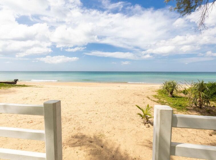 Beach scene seen through a white wooden fence, with sand, calm turquoise water, and a blue sky with clouds far in the distance to the horizon.