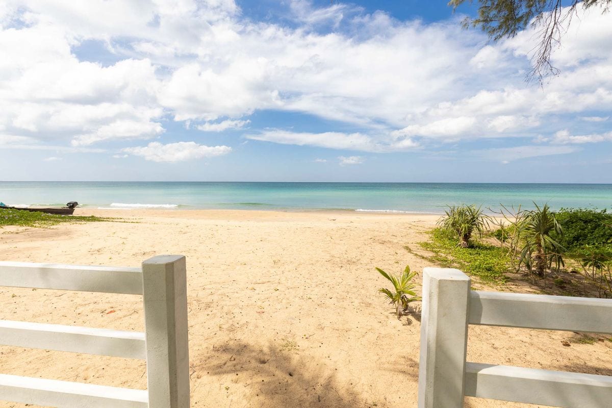 Beach scene seen through a white wooden fence, with sand, calm turquoise water, and a blue sky with clouds far in the distance to the horizon.