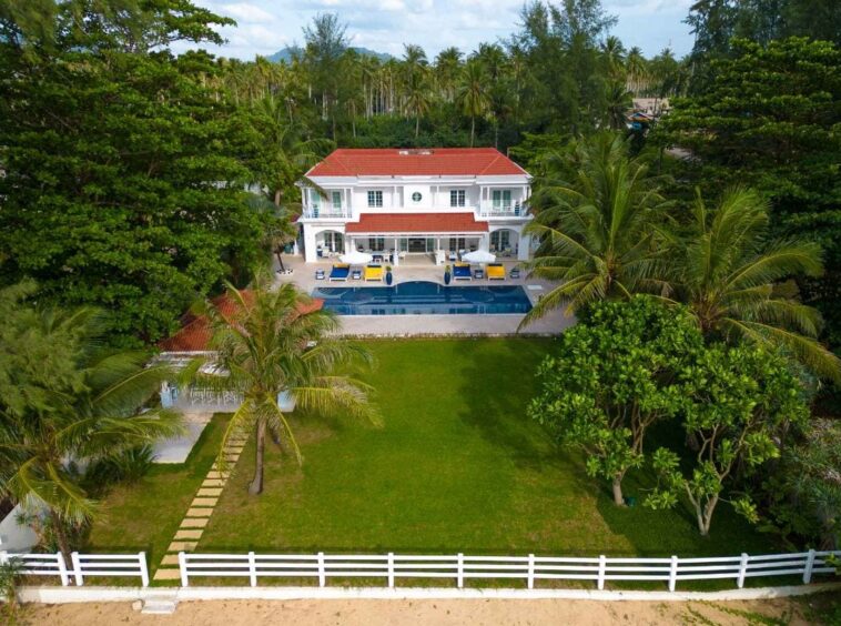 White two-story villa with a red-tiled roof, back porch, and a rectangular pool framed by palm trees and a green lawn.