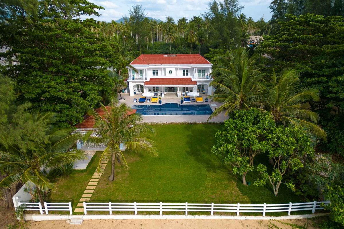 White two-story villa with a red-tiled roof, back porch, and a rectangular pool framed by palm trees and a green lawn.