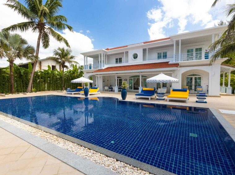 Luxurious white tropical villa with a large blue tiled pool, palm trees, and colorful lounge chairs by the water.