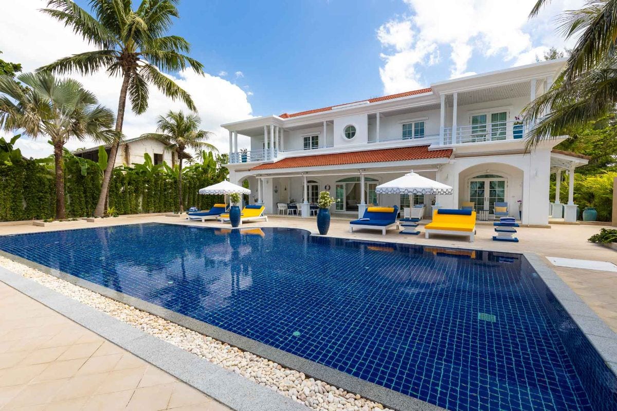Luxurious white tropical villa with a large blue tiled pool, palm trees, and colorful lounge chairs by the water.