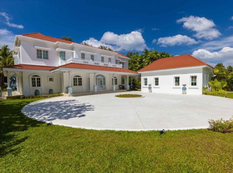 White two-story villa with red-tiled roofs surrounding a circular white driveway and a small landscaped island under a bright blue sky in a tropical setting.